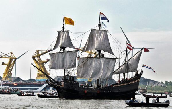 Een groot, historisch zeilschip met drie masten vaart op een rivier of kanaal. De zeilen hangen los en op de masten wapperen Nederlandse en andere vlaggen. In de achtergrond zijn gele hijskranen en windmolens zichtbaar. Er varen meerdere kleine boten vol mensen rondom het schip.